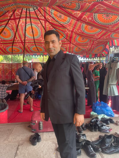 The image captures a bustling market scene in Anajpur, India. A man in a black suit stands prominently, with stacks of black leather shoes arranged to his right. Behind him, a vendor tends to hanging clothes. Other people are visible in the background. The entire scene is covered by a colorful, patterned tent, offering shade.