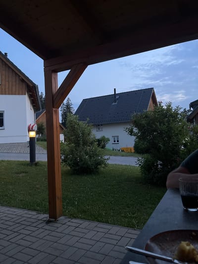 The image shows a view from under a wooden pergola, looking out onto a residential street in Milná, Czechia. The sky is a soft blue with wispy clouds, suggesting it is either early morning or late evening. Two houses are visible, one with a dark gray tiled roof and the other with a lighter-colored tiled roof and white walls. Green trees and grass surround the houses, indicating a well-maintained outdoor space.
In the foreground, the dark brown wooden beams of the pergola create a frame for the scene. To the right, a paved walkway made of rectangular gray bricks leads away from the viewer. A modern black lamppost with a yellow light is positioned along this walkway, casting a warm glow. On the table to the upper right, a person's hand is visible holding a glass, and a plate with food is also present, suggesting a meal or drinks are being enjoyed outdoors. The overall atmosphere is peaceful and relaxed, hinting at a calm evening in a suburban setting. No visible text is present in the image.