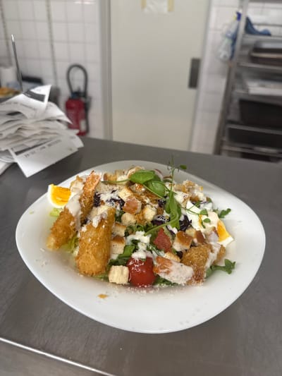 This is a medium shot photograph of a Caesar salad with fried shrimp, served on a white plate. The salad is composed of lettuce, croutons, cherry tomatoes, hard-boiled egg, and shredded cheese, all covered in a creamy dressing. The shrimp is coated in breadcrumbs and fried to a golden brown. The salad is presented on a stainless steel surface, which appears to be a kitchen counter. In the background, there are white tiled walls, a stack of receipts, a red fire extinguisher, and a metal shelving unit with various items. The overall setting suggests a professional kitchen environment, possibly in a restaurant located in Rivesaltes, France, a town known for its wine production and situated in the Occitanie region. There are no people visible in the image.