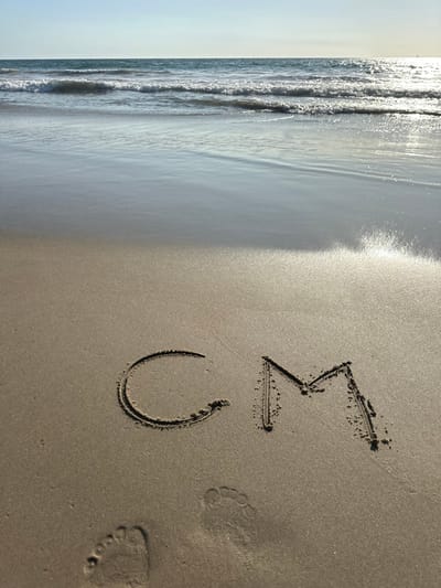 This image depicts a beach scene at Manhattan Beach, United States. The foreground features damp, light brown sand with two distinct human footprints embedded near the bottom left corner. Centrally positioned in the lower half of the frame, the initials "CM" are etched into the sand. The letter "C" is to the left of "M". Beyond the sand, the Pacific Ocean extends into the distance, characterized by multiple parallel waves with white crests breaking and rolling towards the shore. The ocean water exhibits blue and green tones. The horizon is visible, separating the ocean from a clear, light blue sky. Sunlight reflects brightly off the wet sand and the surface of the ocean, especially on the right side of the frame.