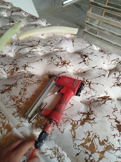 A red pneumatic staple gun lies on a tufted, off-white fabric surface with a gold marbled pattern. Two pieces of curved foam rest atop the fabric. A hand holds an orange air hose connected to the staple gun. In the background is a blurred structure of stacked wooden frames. The fabric surface exhibits recessed, button-like indentations.