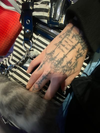 The image depicts a close-up view of a tattooed human left hand resting on a horizontally striped fabric bag, situated in Enghien-les-Bains, France. The hand, with a light skin tone, is positioned palm-down, displaying extensive black ink tattoos across the back and fingers. The tattoo design features intricate, organic patterns resembling thorny vines or stylized tree branches, with some indistinct gothic script visible on the back of the hand near the wrist. The fingernails are short and unpolished.

The hand rests on a bag characterized by alternating black and white stripes, approximately 2.5 cm wide. The bag features black patent leather or synthetic leather accents, including straps, zipper pulls, and trim. Multiple silver-colored metallic zippers with reflective pulls are visible, along with silver-colored hardware such as eyelets, studs, and a D-ring. A small, light gray, fluffy pom-pom charm is attached to the bag via a silver chain.

In the lower-left portion of the frame, a patch of light gray, long-fibered fur or faux fur material is visible, situated beneath the hand and the striped bag. A dark-colored, long-sleeved garment, likely black, is visible on the upper-right, covering the subject's wrist and lower forearm. To the upper-left, a textured, bright red object with a distinct pattern, possibly mesh or woven fabric, is partially visible behind the striped bag. A section of light blue fabric is also visible on the right side, beneath the dark sleeve and bag. The scene is illuminated by artificial lighting, creating a focused foreground against a blurred background.