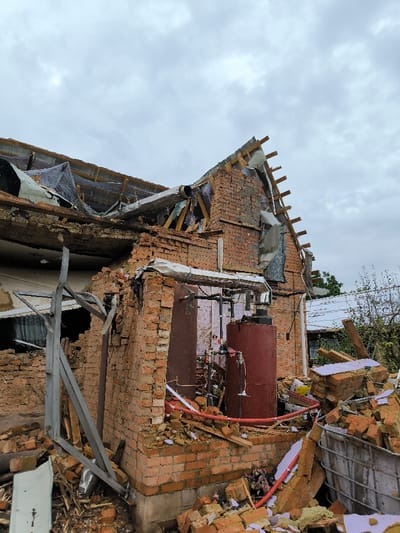 The image depicts a severely damaged brick building in Pysarivka, Ukraine, under an overcast sky. The structure has sustained significant impact, resulting in extensive structural collapse and debris.

The building's roof is largely destroyed, with wooden rafters exposed, fragmented roofing materials (including corrugated sheeting and what appears to be mesh or tarp) hanging precariously. Large sections of the upper brick walls are either missing or heavily fractured, revealing the building's interior. A prominent breach exists in the main brick wall, exposing two large, reddish-brown cylindrical tanks, one taller and one shorter, interconnected by a complex system of black and metallic piping, suggesting a boiler or water heating system. White and red flexible pipes are also visible among the exposed components.

Around the base of the building and extending into the foreground, a substantial accumulation of debris is present. This rubble primarily consists of broken red bricks, shattered insulation material (appearing as purple/pink and yellow fragments), splintered wood, twisted metal elements, and pieces of what resembles shattered glass or plastic. A partially visible, large, white, rectangular corrugated container, possibly an intermediate bulk container (IBC) without its external frame, is filled with rubble on the right side of the foreground. Another less damaged building with a white roof is visible in the background on the right, alongside sparse green vegetation. No individuals or active movements are observed in the scene.