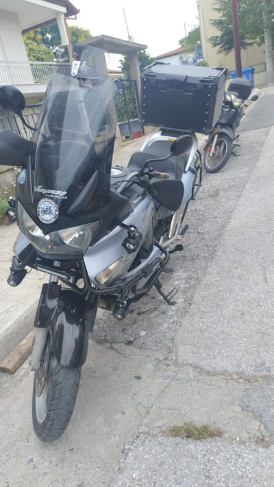 In Νέα Κερασιά, Greece, two motorcycles are parked on a paved street. The primary motorcycle in the foreground is silver and black, with a tall black windscreen, auxiliary lights mounted on the front, and a large black storage box attached to the rear. It has a "Varadero Club Hellas" sticker on the front. A second, smaller black motorcycle is visible behind it, also equipped with a large black storage box. The environment includes a residential building with white railings on the left, a fence, and trees. The ground is a mix of asphalt and cracked concrete, with some sparse vegetation. The sky is overcast.
