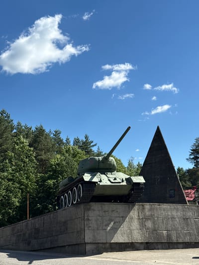 This image, taken in Lenino, Russia, features a monument of a Soviet IS-2 heavy tank on a concrete pedestal. The tank is painted in a camouflage green and is positioned horizontally, facing right. Its main gun is prominently displayed, pointing upwards and slightly to the right. The concrete structure has a rough texture and appears to be part of a larger, angular monument, possibly a memorial.

Behind the tank, a dense canopy of green trees stretches across the frame, suggesting a natural, wooded environment. The sky above is a vibrant blue, punctuated by several fluffy white clouds. The lighting indicates a sunny day, likely midday, casting distinct shadows on the concrete.

The scene appears to be a historical monument or museum dedicated to a military event. There is no visible activity or people in the image. The overall mood is somber and reflective, befitting a war memorial. There is some faint, unreadable text on a dark plaque located on the angular section of the monument. The image is oriented vertically, as if the photographer was looking up at the monument from ground level.