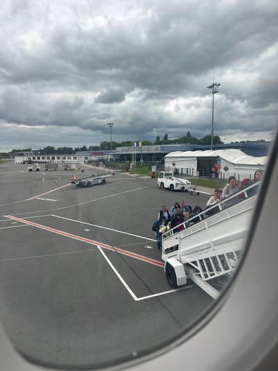 The image captures an airport scene viewed from an aircraft window, partially framed on the right by the window's edge. The sky above is heavily overcast with dark, dense clouds, suggesting gloomy weather conditions.

In the foreground, a white mobile passenger staircase is positioned next to the aircraft, filled with people, likely passengers, who appear to be either boarding or disembarking. Some individuals are standing on the steps, while others are seated on platforms along the staircase.

The tarmac below is asphalt, clearly marked with white and red lines characteristic of airport ground operations. Several ground support vehicles are visible: a white pushback tug is situated near the passenger staircase, and further down the apron, other service vehicles, including a long, low vehicle possibly used for baggage or cargo, can be seen. A person wearing a high-visibility vest is discernible near one of these vehicles, indicating airport staff.

In the background, several airport buildings are visible, including large white tent-like structures, which may be terminal extensions or hangars, and other smaller utility buildings. A tall pole topped with lights or antennae stands prominently against the cloudy sky. Near the buildings, flags on poles are faintly visible, with one resembling the European Union flag. Visible text includes a red "STOP" sign and a blue sign with a white arrow near the buildings, and ground markings like "2" and "3" on the tarmac.

The scene depicts a typical airport operation, likely a passenger transfer, during daylight hours under an overcast sky. The location context is Tillé, France.