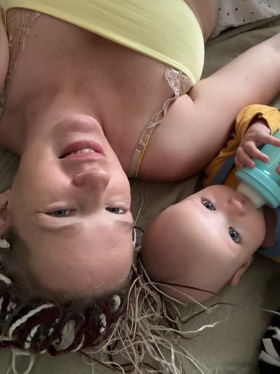The image shows a close-up of a woman and a baby lying down, with their heads angled towards the camera. The woman, wearing a yellow top with lace trim, is positioned on the left side of the frame, and her face is visible upside down. She has light-colored eyes and pink lipstick. The baby, on the right side of the frame, is also upside down and is holding and drinking from a turquoise baby bottle. The baby has light-colored eyes and is wearing a yellow long-sleeved shirt. Their hair appears to be a mix of blonde and brown, with some lighter streaks. They are lying on a textured surface, possibly a bed or couch, which appears to be a muted green color. The overall lighting suggests an indoor setting. There is no indication of the city of Бордуки, Russia in the image.