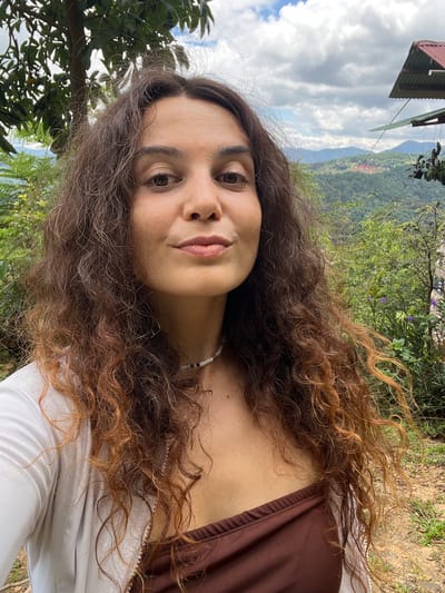 A medium shot portrait of a woman with long, curly brown hair, wearing a white open shirt over a brown top, is captured outdoors. She has fair skin and is looking towards the camera with a neutral expression. A pearl necklace is visible around her neck. The background reveals a lush green landscape with rolling hills and trees, under a cloudy sky. A portion of a building with a red roof is visible in the upper right corner, suggesting a rural or mountainous setting, possibly in the city of Sơn Điền, Vietnam. The ground in the foreground is covered with dirt and small patches of grass.