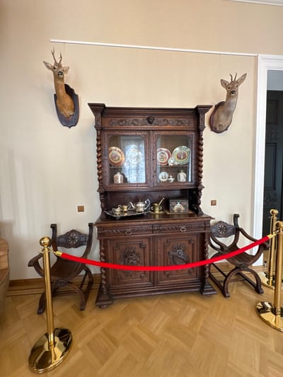 A photograph shows the interior of a building in Добасненскі сельскі Савет, Belarus. On the beige wall, a wooden cabinet with glass doors displays various objects. Above and below the cabinet are taxidermied deer heads. A red velvet rope, supported by golden stanchions, is placed to keep people away from the cabinet. Two wooden chairs stand beside the cabinet. The floor is wooden, and the scene is well-lit.