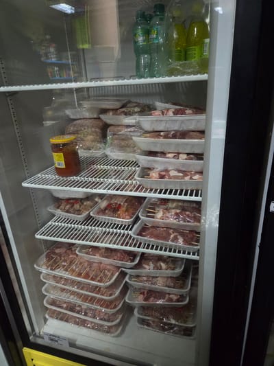 The image shows the inside of a refrigerated display case, likely in a store located in Pripek, Bulgaria. On the left side of the case are bottles of liquid, possibly drinks. The main section of the case is filled with packaged skewers of meat. These are arranged in plastic containers and hung on a wire rack system. Near the bottom, a jar of yellow-labeled food product is visible. The lighting is bright, typical of a retail refrigerator.