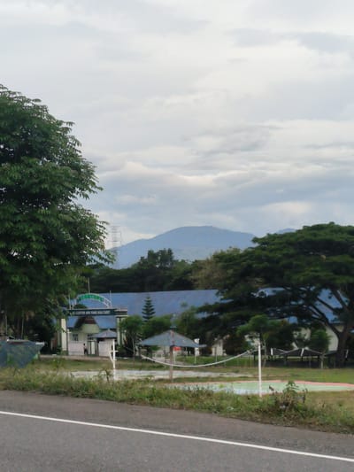 The image presents a roadside view of a landscape in Meunasahbaro, Indonesia, under an overcast sky. In the foreground, an asphalt road with a solid white line on its right edge leads towards the middle distance. Green grass and shrubs line the road, transitioning into an open field area that features a marked court with a white net, resembling a volleyball court.
In the mid-ground, a building complex with blue roofs is prominent, characterized by an archway at its entrance. The archway prominently displays the text "KE GONTOR APA YANG KAU CARI?" (To Gontor, what are you looking for?). A lone individual is visible standing near the entrance of this building. The scene is framed by large, leafy green trees on both the left and right sides, and smaller vegetation is scattered throughout.
Further in the background, a range of mountains rises, appearing hazy against the sky. A power transmission tower is distinctly visible on the left, silhouetted against the distant mountains. The sky is largely covered with grey, diffused clouds, indicating an overcast day with soft, non-directional lighting. The overall atmosphere appears tranquil, depicting a serene, possibly semi-rural, setting that could be associated with an educational or institutional compound, given the prominent sign.