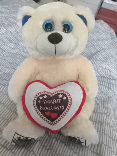 A cream-colored teddy bear with bright blue eyes and blue inner ears sits on a grey, patterned surface. The bear is holding a heart-shaped pillow in its paws. The pillow is white with a red border and features a brown gingerbread-style design with the words "VOLKSFEST BREMERHAVEN" and two red hearts. The location is unknown, but the pillow indicates a connection to the Volksfest in Bremerhaven, Germany.
