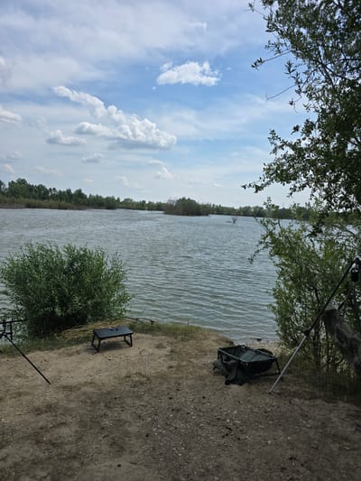 The image depicts a serene lakeside scene in Krčmaň, Czechia, under a partly cloudy sky. In the foreground, a sandy bank slopes down to the water's edge, with sparse patches of grass and gravel. To the left, two fishing rod rests are planted in the ground, and a small, black portable table sits on the bank. A bundle of yellow fishing line is draped over the table. In the center, a green fishing weigh sling is placed on the ground, with a silver pole propped up behind it, partially obscured by foliage. The lake itself is a calm expanse of water, with gentle ripples across its surface. The far shore is lined with a dense band of green trees, their foliage creating a natural backdrop. The sky above is a mix of blue and white, with scattered cumulus clouds adding depth and texture to the scene. The overall impression is one of tranquility and a quiet fishing spot.