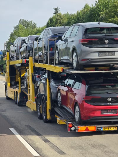 A yellow car carrier truck is driving on a highway with several Volkswagen ID.3 electric cars loaded on its upper and lower decks. The truck is in the right lane, with trees and foliage visible in the background. The sky is overcast. The license plate of the truck is visible as 59-WR-NZ. The scene appears to be on a highway near Oberselters, Germany, based on the presence of German license plates on the cars and the environment.