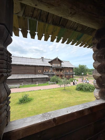 The image shows a view from a wooden porch through decorative carved eaves of a wooden building. In the foreground, the textured, aged wood of the porch structure is visible on the left and right edges, framing the scene. A weathered wooden railing runs across the bottom of the frame. Beyond the porch, a large, multi-story wooden building with a gabled roof, numerous windows, and balconies is the central subject. A pathway made of gravel and small stones leads to the building, bordered by a neatly mowed green lawn. In the middle ground, several people are walking on the gravel path in front of the wooden building. Some are gathered near a lamppost. The background consists of lush green trees and a cloudy sky. The location is identified as Nevskiy Parkleskhoz, Russia.