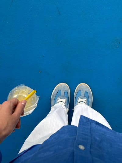 A top-down, medium shot shows a person standing on a blue surface in Tambon Sa Khu, Thailand. The person wears blue sneakers with white laces, white pants, and a blue shirt. The person holds a clear plastic cup with a yellow straw. The background is a solid blue surface.