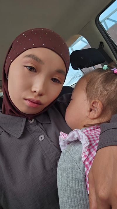 In Kyzyl-Suu, Kyrgyzstan, a young woman wearing a maroon hijab adorned with rhinestones, and a grey button-up shirt, is holding a sleeping baby. The baby, with its eyes closed and cheek pressed against the woman's chest, is wearing a grey ribbed long-sleeve shirt and a pink and white checkered bib. The scene appears to be inside a car, with the car's interior visible in the background, including a headrest and a window.