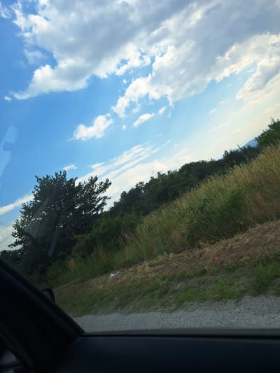 This media file depicts a daytime scene with a clear blue sky and scattered white clouds. The perspective suggests the image was taken from inside a vehicle, looking out. The scene transitions from a bright sky on the left to a grassy roadside with trees on the right, bordering a gravel path. The vegetation appears dry in some areas and green in others, with a prominent large tree in the foreground on the left side of the frame. There are no people visible. The lighting and cloud formations suggest it is daytime, likely late morning or afternoon, with fair weather conditions. No text is visible in the image. The overall impression is that of a scenic drive through a rural or semi-rural environment in Bulgaria.