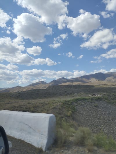 The image depicts a landscape view of rolling hills and mountains under a partly cloudy blue sky. In the foreground, a white concrete barrier is visible on the left side of the frame. The ground appears to be covered with dry vegetation and gravel. Several utility poles and wires stretch across the middle ground. The mountains in the background are barren and arid, with visible shadows indicating the direction of the sun. The scene suggests a rural or mountainous environment.
