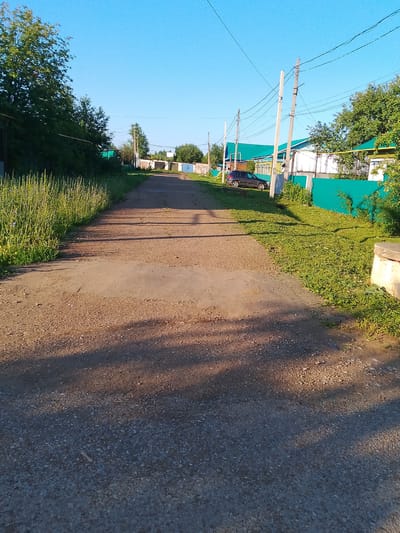 The image depicts a rural road in Kranokholmskiy, Russia. The road is unpaved, made of gravel and dirt, and appears to be a residential street given the presence of houses and fences lining its sides. Tall green grass and trees grow along the left side of the road, while on the right, manicured grass borders a bright green fence. Several utility poles with numerous wires stretch across the frame overhead, indicating a connection to electricity or telecommunications. A dark-colored car is parked on the side of the road near one of the houses, which has a green roof. The sky is a clear, bright blue, suggesting a sunny day. The lighting indicates that the sun is low, casting long shadows across the road, implying it is either early morning or late afternoon. There are no people visible in the image. The scene is one of a quiet, possibly remote, residential area.