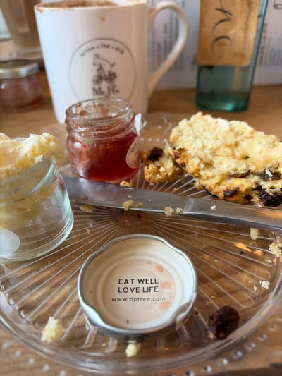 The image presents an overhead, close-up view of a partially consumed cream tea service on a wooden table at Maison H cafe in Ambleside, United Kingdom. On a clear, ridged plastic serving plate, a silver-colored metal lid from a Tiptree product is inverted, displaying the text "EAT WELL LOVE LIFE" and "www.tiptree.com". A metal butter knife rests diagonally across the plate, coated with scone crumbs. To the left, a small clear glass jar contains a substantial amount of pale yellow clotted cream, with some cream residue on its rim. Another small clear glass jar, approximately half-full, holds red jam. Two substantial pieces of a raisin scone, exhibiting a golden crust and interior, are positioned on the right side of the plate, surrounded by scattered crumbs.

In the midground, a white ceramic mug, identified by a circular 'M.H.' logo depicting a cyclist and 'FELLOW + THY + BIRD' text, contains a foamy, dark beverage, consistent with coffee or hot chocolate. A smaller lidded jar is partially visible behind the clotted cream jar. To the right, a tall, turquoise-tinted glass bottle with a brown paper tag bearing handwritten script stands partially obscured. The background elements are softly blurred, confirming an indoor cafe setting. No individuals are visible, but the arrangement implies recent dining activity.