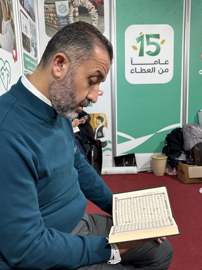 This image depicts an indoor scene in the city of Al-Ghizlaniya, Syria, showing a man seated, focused on an open book. The man, with dark hair and a short, gray beard, wears a teal-blue long-sleeved sweater over a white collared shirt and dark gray trousers. He is holding a hardbound book, open to pages displaying Arabic script, and appears to be reading or reciting. A white wristwatch is visible on his left wrist. The environment features a red carpeted floor. In the background, a large green and white banner displays the number "15" with leaf designs and Arabic text, which translates to "15 years of giving." To the left, a portion of another banner with images is visible. Additional background elements include a white waste bin, a brown cardboard box, miscellaneous bags or clothing, and a black folder or case. Indistinct figures and a cutout image of a man are also visible further in the background.