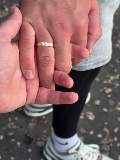 A close-up, over-the-shoulder shot shows two pairs of hands clasped together, with a gold band visible on one of the ring fingers. The background suggests an outdoor urban environment, possibly a sidewalk or paved area with scattered debris. The hands appear to belong to individuals wearing athletic attire, including white socks with a black swoosh logo and athletic shoes. The overall lighting indicates daytime, with a slight warmth to the skin tones. The scene is devoid of any specific landmarks that would pinpoint its location within Santa Cruz de Tenerife, Spain.