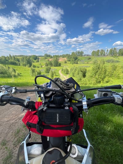 The image depicts a first-person perspective from atop a motorcycle, stationary on a dirt path in Новочувашский, Russia. The motorcycle's black handlebars, silver clamps, and control levers are prominent in the lower foreground. A pair of grey and blue gloves, marked with "100%", rests on the handlebars. Below the handlebars, a red fabric bag is attached, and a black electronic device or case, labeled "panaride", is mounted above it. A portion of the motorcycle's light-colored fuel tank is visible, featuring a fuel cap with the inscription "DO NOT OPEN HOT".

The scene unfolds into a rural landscape characterized by rolling green hills. A winding dirt track extends into the midground, flanked by lush green grass and numerous deciduous trees with dense foliage. Patches of purple flora are visible within the grassy areas on the hillsides. In the distance, several light-colored buildings, presumed to be houses, are situated on a ridge, partially obscured by trees. To the left, a distinct area of disturbed ground or a quarry with lighter soil is visible on a hillside. The sky above is a clear blue, dotted with scattered white cumulus clouds. The immediate ground beneath the motorcycle is a mix of dirt and sparse green grass, transitioning to thicker grass on the right.