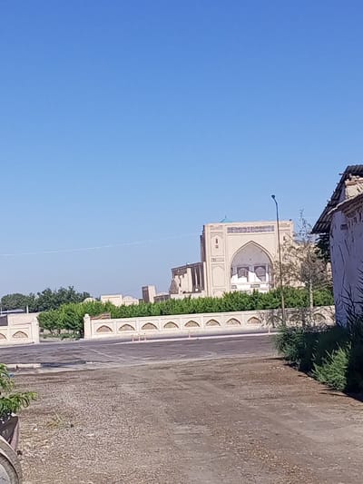 A clear, blue sky looms over an outdoor scene in Shar-Bakr, Uzbekistan. In the background, a large, beige, arched building with a turquoise dome dominates the landscape. A decorative wall with repeating arched cutouts runs across the middle ground, separating the building from a paved area in the foreground. Green trees and bushes line the decorative wall. A single street lamp stands near the central building. To the right, a portion of a white building with a dark, corrugated roof is visible. The foreground consists of a textured, gravelly ground. The image appears to be a daytime shot, possibly taken from a street or parking area.
