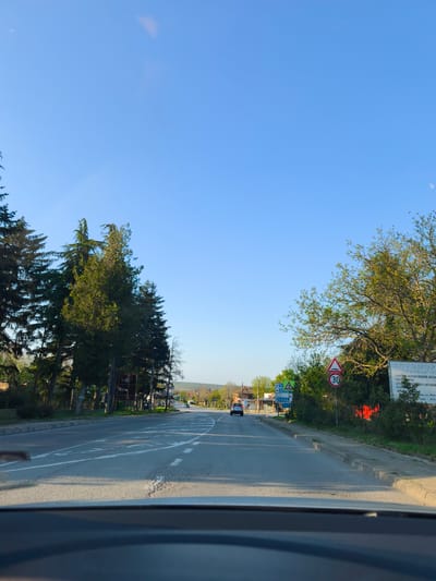 The image depicts a straight road in Staro Oriahovo, Bulgaria, viewed from the interior of a vehicle, with the dashboard visible at the bottom. The road, featuring white markings including a solid line on the left and dashed lines on the right, appears to lead into a settlement. On both sides, rows of trees line the road, with more dense coniferous trees on the left and deciduous trees with fresh spring foliage on the right.
The scene is set on a clear, sunny day, indicated by the bright blue sky and distinct shadows cast by the trees and a car on the road. Sunlight illuminates the right side of the road and trees, suggesting it might be morning or late afternoon.
In the distance, a blue car is visible on the road, with another darker vehicle further ahead. Several road signs are present on the right side. A prominent red-bordered triangular sign indicates a hazard, and directly below it is a circular speed limit sign displaying "30", suggesting a 30 km/h speed limit. Further along the road on the right, a large billboard is erected.
Visible text from the billboard includes Cyrillic script: "АВТОМОР" (AUTOMOR), "ТУЗАРИ 20", "ИЗКУПУВА ВСИЧКИ ВИДОВЕ АВТОМ..." (BUYS ALL TYPES OF AUTOM...), "ЧАСТИМИКРАП", "ПРОДАЖБИ НА НОВИ И АВТОМОБ..." (SALES OF NEW AND AUTOMOB...), "УПОТ" (USED), "ово, ул. Лом" (ovo, Lom Str.), and "преди лук" (before Luk...). Smaller, less legible directional signs are also seen on the right, next to the road. The overall impression is a calm drive through a populated, possibly rural-urban fringe area.