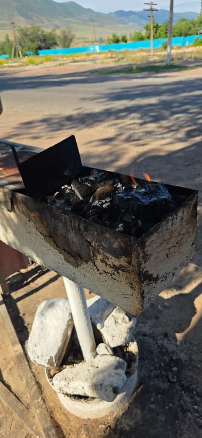 In Sasyk-Bulak, Kyrgyzstan, a close-up shot focuses on a rustic, weathered metal brazier filled with glowing charcoal and small flames. The brazier, positioned outdoors, appears to be set on a concrete base with several large, irregular concrete chunks. A black metal flap is angled within the brazier, partially obscuring the embers. The background shows a paved road with visible tire tracks and shadows cast by an unseen object, suggesting a sunny day. Beyond the road, a vibrant turquoise wall runs along the landscape, backed by dry grass and trees. In the distance, rolling hills form a natural backdrop under a clear sky. A utility pole with wires is also visible. The overall scene depicts an outdoor setting, possibly a roadside or park area, with a focus on the functional, yet aged, cooking equipment.