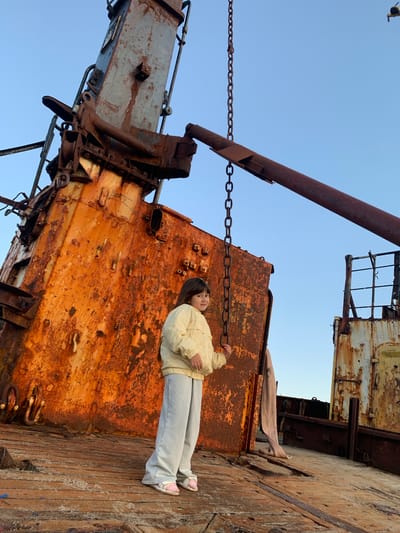 A young girl stands on the deck of a rusty shipwreck in Teriberka, Russia. The girl, wearing a light yellow jacket and light blue pants, is looking towards the camera. She has long, dark hair. Behind her is a massive, heavily rusted crane structure that dominates the left side of the frame. Chains hang from the crane. The deck beneath her feet is made of weathered wooden planks. The background is a clear blue sky. Another rusted ship structure is visible to the right. The overall scene depicts the decay and abandonment of industrial equipment in a coastal environment.