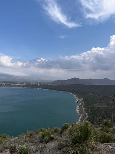 The image presents a vertically oriented landscape of Alchichica, Mexico. The view is dominated by a large body of deep blue water, suggestive of the Alchichica crater lake. The shoreline is rugged with vegetation extending to the water's edge. On the right, a rocky cliff is covered with agave-like plants. A narrow road traces along the rocky shoreline. In the distance, the city Alchichica lies in front of mountains obscured by a haze, with cloud formations above a blue sky to the left.