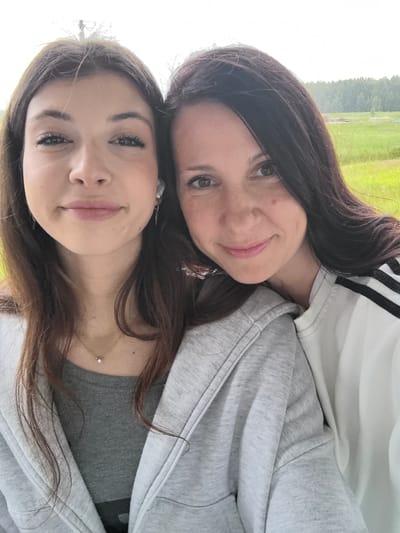 The image is a close-up selfie of two individuals, likely mother and daughter, outdoors with a blurred background of greenery and trees, possibly in the city of Usady, Russia. The person on the left is younger with long brown hair, wearing a grey hooded jacket over a grey t-shirt and a delicate necklace. She has on white earbuds. The person on the right has long dark brown hair and is wearing a white sweatshirt with black stripes on the sleeve. Both are looking at the camera with gentle smiles. The lighting suggests it is daytime, possibly overcast.