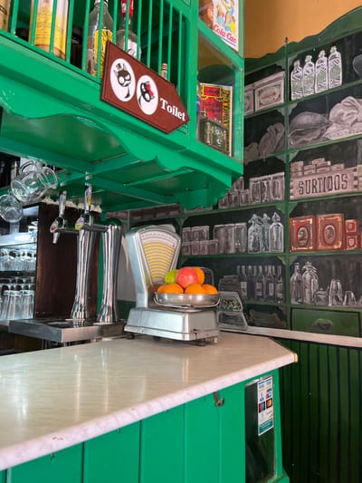 The image shows a bar counter with a green tiled surface. There are three taps for dispensing beer, and a scale for weighing produce sits on the counter. Behind the counter, there are several bottles and jars on shelves and a green sign that reads "Toilet" with cartoon cats on it. The bar counter has a green tiled exterior. The scene is located at the coordinates 27.907, -15.447, which is in the town of Agüimes, on the island of Gran Canaria, Spain.