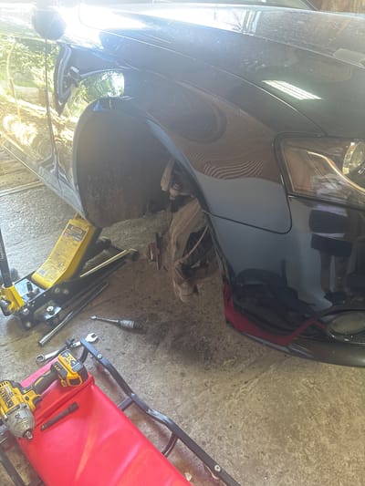 This image depicts an automotive repair scene, likely in Bivolare, Bulgaria. A black car, possibly an Audi, is elevated by a yellow hydraulic jack, with its front passenger-side wheel removed, exposing the brake assembly. Tools, including a DeWalt impact wrench and various sockets, are scattered on the concrete ground, some resting on a red mechanic's creeper. The environment appears to be a garage or workshop, with sunlight filtering in and reflecting off the car's surface, highlighting dust and dirt. The scene suggests a do-it-yourself car maintenance operation.