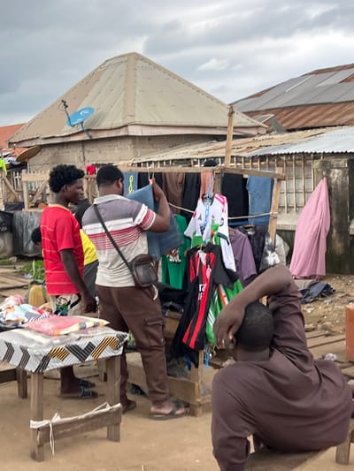 In Oko Amakom, Nigeria, an outdoor market scene unfolds under a cloudy sky. Three men are present. One man, wearing a red striped t-shirt, stands to the left, facing right. Another man, in a striped grey, red, and white t-shirt and brown cargo pants, stands in the center, holding up a pair of blue denim jeans. He has a dark shoulder bag. A third man, wearing a dark brown robe, is seated on a low wooden stool at the right, facing away from the camera, with his hand on his head.
In the foreground, a small wooden table covered with a patterned cloth displays folded items wrapped in clear plastic. Behind the people, a makeshift stall displays an assortment of clothing, including shirts and jerseys, hanging on a wooden frame. Various types of roofs, some made of corrugated metal and others with solar panels, are visible in the background, along with simple building structures. The ground is dirt.