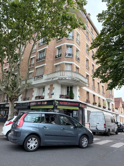 This is an eye-level, medium shot of a street scene in Maisons-Alfort, France. The main subject is a multi-story brick building with white trim and balconies. In front of the building, several cars are parked along the street. A gray minivan is in the foreground, obscuring a portion of a crosswalk. To the left of the minivan, a white car is parked. Behind the gray minivan, a white van is parked, and to its right, a dark-colored car is parked. The building has several storefronts on the ground floor, including one with a sign that reads "BARBER OP" and another that says "SAVEURS DES ILES". Trees with green foliage are visible throughout the frame, partially obscuring the upper floors of the building and the sky. The day appears overcast.
