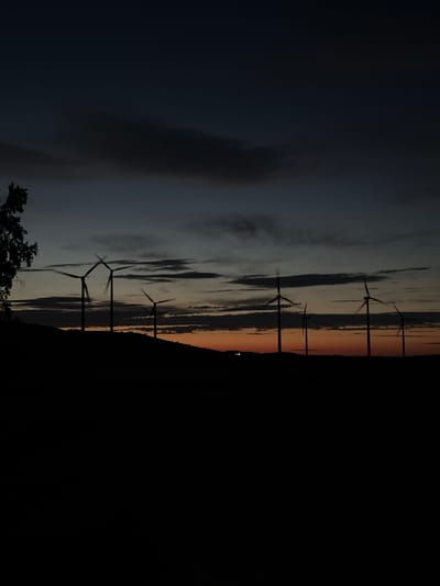 The image depicts a silhouetted landscape at dusk or dawn, with several wind turbines visible on a ridge. The sky is a gradient of dark blue to a faint orange near the horizon, with some cloud streaks. A dark silhouette of trees is on the left side of the frame, and the ground is also in silhouette. The wind turbines have their blades blurred, suggesting they are in motion. Two small, bright lights are visible in the distance on the ground, possibly from a building or vehicles. The scene is likely an outdoor, rural setting. The location of Bereznyakovka, Kazakhstan is not explicitly discernible from the visual information provided in the image itself.