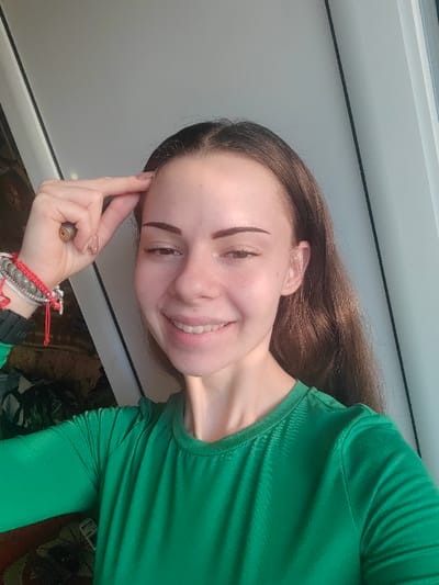 In a selfie-style photograph taken in Dolna Kremena, Bulgaria, a light-skinned female with long brown hair parted in the middle is positioned centrally, facing the camera directly. She smiles, revealing teeth. Her right hand is raised, with her index finger and thumb gently pinching the skin near her right temple. A ring is visible on her right ring finger, and her fingernails are painted a light nude color. On her left wrist, she wears multiple accessories, including a dark-colored wristband, several red string bracelets, and a multi-strand beaded bracelet in shades of grey and white. She is wearing a long-sleeved, crew-neck green shirt with a subtle textured pattern. The background is partially visible; to the left, a dark, indistinct area with hints of vegetation or patterned fabric is present, while to the right, a bright, light-colored vertical surface, possibly a window frame or wall, is illuminated. Natural light appears to originate from the right side of the frame.