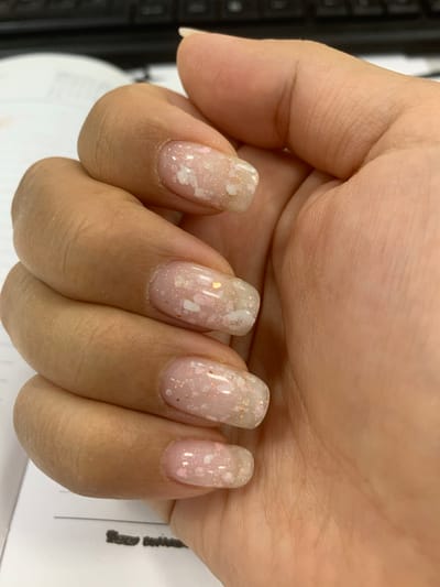A close-up, top-down view shows a person's left hand with manicured fingernails resting on a white surface, possibly a desk with papers. The nails are adorned with a light pink base coat and adorned with small, irregular white and iridescent flakes, as well as subtle gold glitter. The hand is positioned as if it is holding or about to pick something up. The background is blurred, but appears to be a dark keyboard and a white document. The location in Urtaaul, Uzbekistan is not discernible from the image.