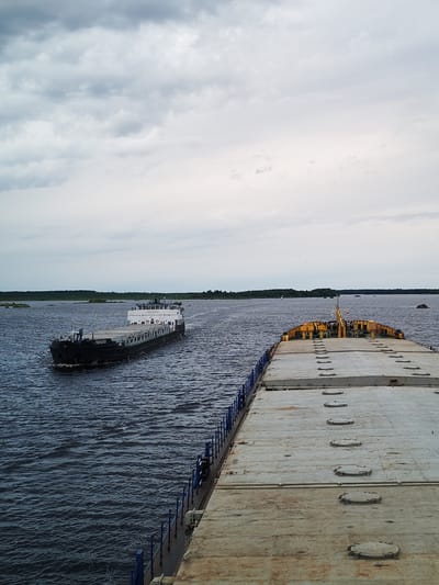 The image depicts a wide, dark blue body of water under a cloudy, grey sky. In the distance, a treeline forms the horizon. Two barges are visible on the water. The closest barge, in the foreground, is large and flat with a grey deck featuring multiple circular hatches. It is positioned on the right side of the image, facing away from the viewer, with its left side extending out of frame. Blue railings line the side of this barge. Further in the distance, on the left side of the image, a second, smaller barge is moving towards the viewer. This barge has a black hull and a white superstructure. Text, which appears to be Russian, is visible on the hull and superstructure of this second barge. The water is choppy, with visible waves. The overall scene suggests a commercial waterway or river. The location is identified as Novokemskii, Russia.
