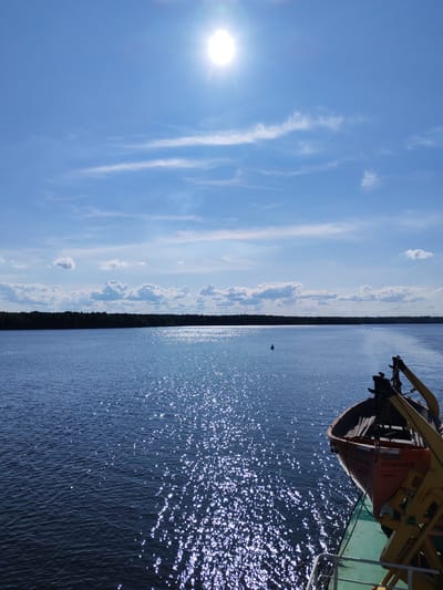 This image depicts a sunny day on a body of water, likely a river, with a dark, forested shoreline in the distance. The sun is high in the sky, casting a bright glare on the water, which is a deep blue and flecked with sparkling highlights. Several white clouds are scattered across the bright blue sky. In the foreground, the edge of a vessel is visible, with a life raft hanging from a davit. The life raft is orange and appears to be made of fiberglass. Text is visible on the side of the life raft, though it is partially obscured and difficult to read clearly. The scene suggests a calm and pleasant day for a boat trip or transit in the Shaima, Russia region. The time of day is midday, given the high sun. The overall mood is serene and tranquil, characteristic of a beautiful summer day on the water.