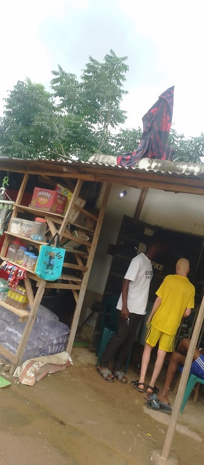 The image depicts an outdoor scene in Eka, Nigeria, likely in front of a small shop. A wooden structure, appearing to be a makeshift store, is visible, with shelves filled with various goods including bottled drinks, packaged items like "Mimee" biscuits, and clear plastic containers with contents. A blue tote bag with a graphic hangs on one of the shelves.

Two men are present in the foreground. One man, wearing a white t-shirt and dark pants, stands facing away from the camera, holding a mobile phone. The other man, who appears to have albinism, is dressed in a bright yellow t-shirt and shorts and stands with his back to the camera, facing the same direction as the first man. Another person's legs are visible further to the right, sitting on a chair.

The ground appears to be dirt or mud, suggesting recent rain or a generally unpaved area. In the background, trees and foliage are visible against an overcast sky, with a dark fabric, possibly a blanket or flag with red and black patterns, draped over the corrugated metal roof of the structure. A single light bulb hangs from the ceiling of the shop. The overall scene suggests a casual, everyday setting within a Nigerian community.