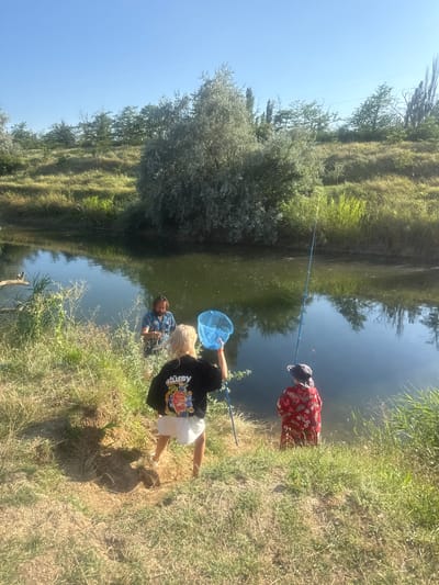 In the city of Pushkinskoye village settlement, Ukraine, on a sunny day, three individuals are engaged in activities by a body of water. A man with a beard, wearing a patterned short-sleeved shirt, stands in the shallow water near the grassy bank. A young person with blonde hair, facing away from the camera, wears a black sweatshirt with "stüssy" printed on it and white shorts, holding a blue fishing net. To their right, another young person, also facing away, wears a red patterned shirt and a dark hat, holding a blue fishing rod. The water's surface reflects the sky and surrounding greenery. The environment features a sunlit grassy bank with dry patches and various vegetation, leading to the tranquil water.