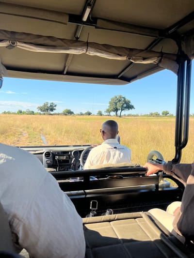 The image shows the interior of a safari vehicle in Kakoro, Botswana, with a driver wearing a "Nakwa Safaris" shirt and glasses visible at the steering wheel. The vehicle is open-topped with a canvas roof support structure. Outside the vehicle, there is a flat, grassy landscape with scattered trees under a clear blue sky. A passenger is partially visible to the driver's right, wearing a light-colored shirt and possibly a hat. The vehicle's dashboard and various controls are in view, and there's a seat behind the driver. The angle indicates that the photo was likely taken from the backseat.