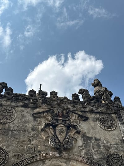 The image shows a close-up, low-angle view of the facade of the former Convent of San Andrés Apóstol in San Andrés Calpan, Mexico. The ancient stone facade is weathered and shows intricate carvings. At the top of the frame, a bright blue sky is visible with a few white clouds.

The facade features a central relief of a winged figure wearing a crown, with a lion's mane or beard. The figure is seated and appears to be holding a shield or crest. To the left and right of this central figure are circular medallions with decorative elements and what appears to be Latin text. On the upper edges of the facade, several stone finials are visible, some resembling small crowns, and a sculpted figure of a person in a praying or contemplative pose is perched on the right. The stonework is textured and shows signs of age. The overall impression is one of historical grandeur and weathered beauty.