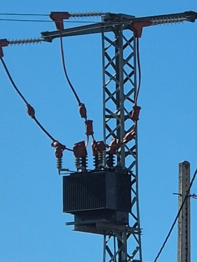 The image depicts a segment of an electrical power transmission structure against a clear blue sky. The structure is metallic and lattice-like, supporting high-voltage electrical components. These include a black, box-shaped transformer, insulators, and electrical wires. The red insulators are spaced along the wires and at the connection points to the transformer. The location of the scene is Collado Del Mirón, Spain.