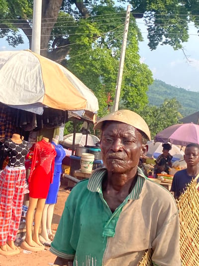 This image depicts an outdoor market scene in Amaseri, Nigeria, under bright daylight. In the foreground, an older man with dark skin and a weathered face, wearing a two-toned polo shirt (green on the left, tan on the right) and a light-colored bowl-shaped object on his head, looks directly forward. A woven, light-colored structure is partially visible to his right.
To the left of the man, a market stall is covered by a large umbrella with alternating white and tan segments. Beneath this umbrella, three mannequins are displayed: one wears a black and white patterned top with red and white plaid pants, another wears a long-sleeved red dress with sequined details, and a third wears a blue dress. Racks of clothing are visible behind the mannequins. A white plastic bucket labeled "ACLUX" rests on a surface near the mannequins.
In the midground to the right, a young boy in a dark t-shirt is partially visible, looking forward. Behind him and further back, multiple other market stalls are indicated by various umbrellas, including a prominent purple one. People are visible behind these stalls, with merchandise displayed on tables.
The background features a tall, light-colored utility pole with multiple wires extending across the frame. Dense green trees are present throughout the background, and rolling green hills are visible under a light blue sky on the far right. The ground surface appears to be dirt or sand.