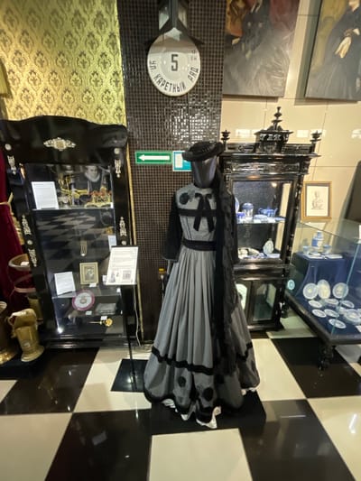 The image depicts a museum exhibit, likely in Voskhod, Russia, showcasing historical artifacts. In the foreground, a mannequin is dressed in a grey and black vintage dress with a matching hat and scarf. To the left of the mannequin is a black display cabinet filled with various items, including framed pictures and what appear to be jewelry and decorative objects. To the right of the mannequin, another black display cabinet contains blue and white porcelain items. Behind the mannequin, a tiled wall features a round sign with the number "5" and Russian text, along with a green directional arrow. The floor is a black and white checkered pattern. Paintings of historical figures or scenes are visible on the wall in the background, and the overall lighting suggests an indoor setting. There are no identifiable people actively engaged in the scene, only the mannequin and displayed items.
